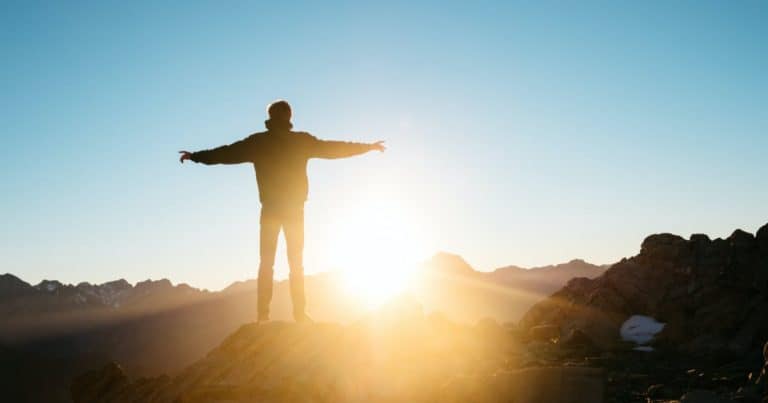 Man on mountain top with outstretched arms looking at the sun on the horizon.