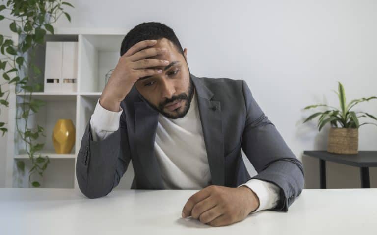 A man in a suit sitting at a desk with his hand on his head looking frustrated.