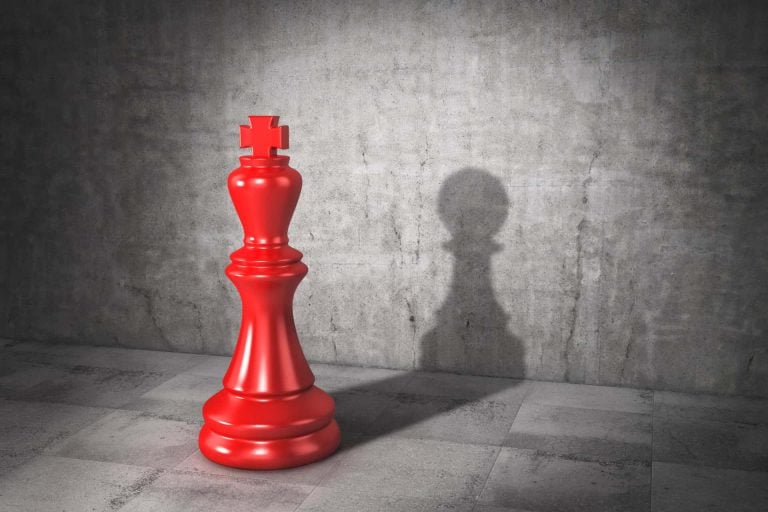 A large red chess king stands on a tiled floor casting a pawn shadow against a gray concrete wall.