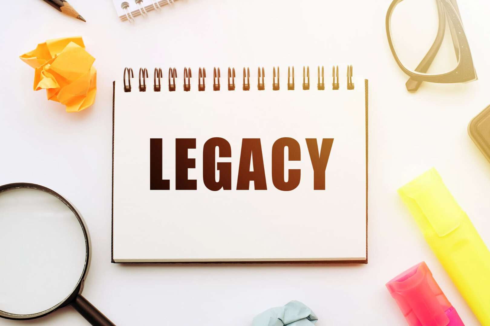 A spiral notebook with the word "LEGACY" in bold letters, surrounded by office supplies like a magnifying glass, highlighters, glasses, and crumpled paper.