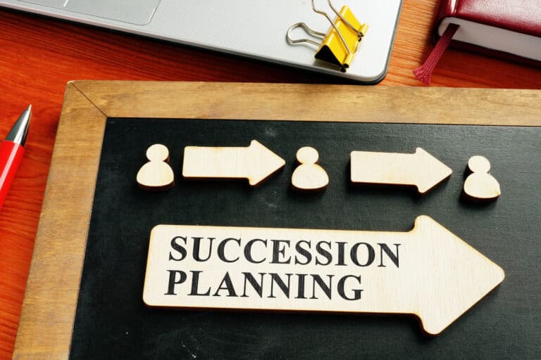 Chalkboard with wooden pieces shaped like people and arrows, and a text sign that reads "Succession Planning." A pen, a binder clip, and part of a laptop are visible on a wooden surface.