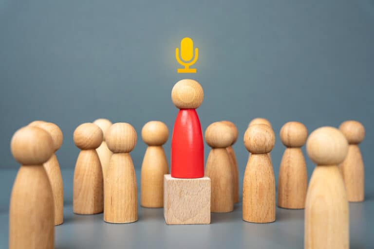 Wooden figures surround a central red figure on a pedestal, symbolizing leadership or speaking. A yellow microphone icon is above the red figure's head.