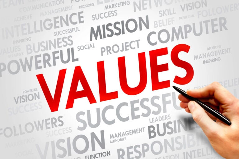 A hand holds a pen near the word "VALUES" in large red text, surrounded by gray business-related words like "MISSION," "SUCCESSFUL," and "INTELLIGENCE" on a white background.