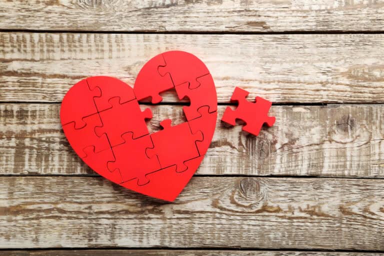 A red heart-shaped jigsaw puzzle with one piece missing, placed on a rustic wooden surface. The missing piece is positioned nearby.