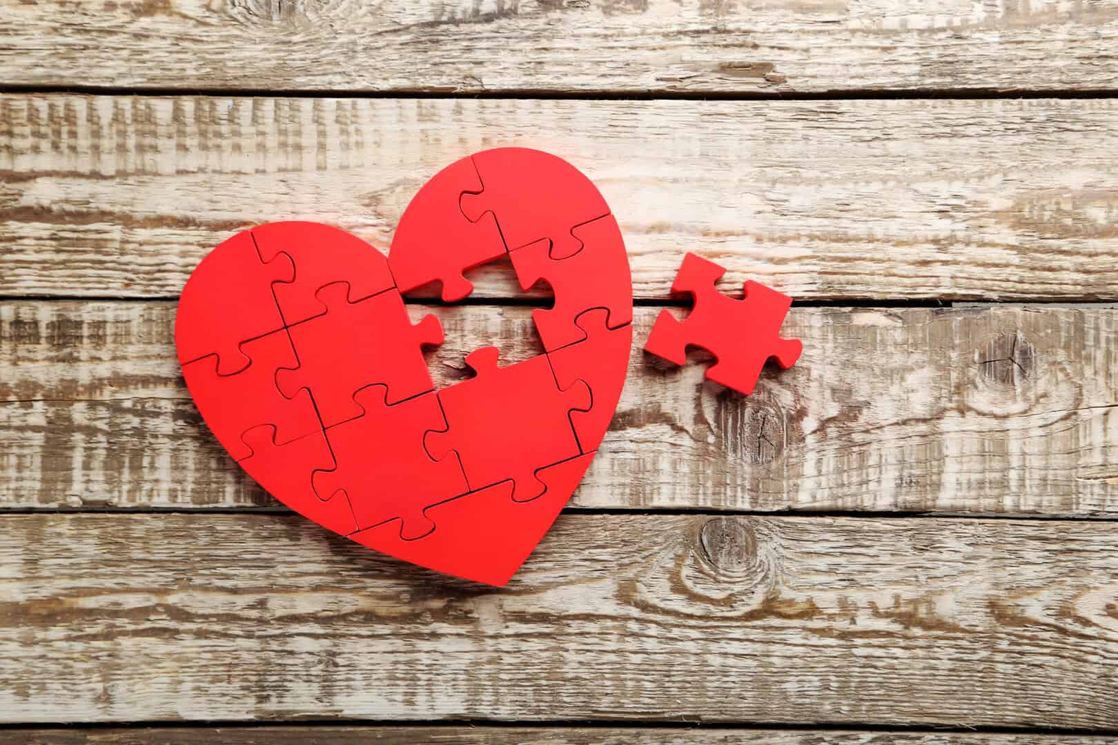 A red heart-shaped jigsaw puzzle with one piece missing, placed on a rustic wooden surface. The missing piece is positioned nearby.