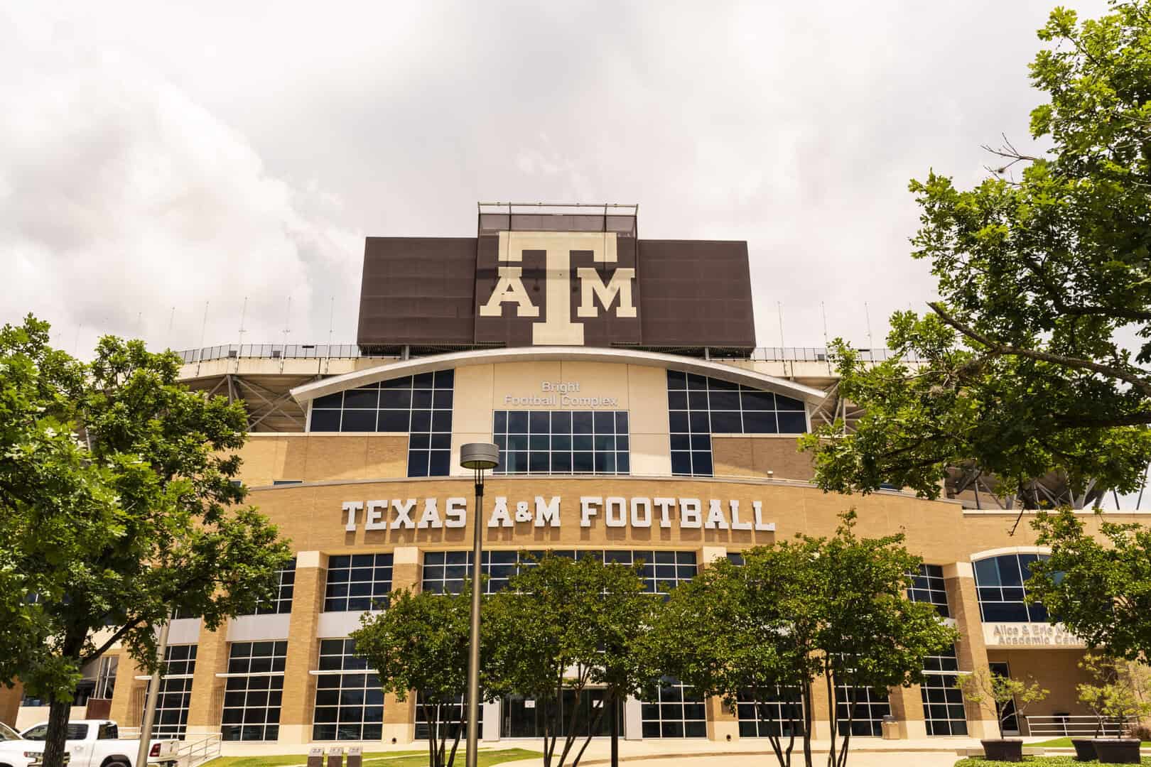 Exterior view of the Texas A&M Football stadium, featuring a large "ATM" logo and surrounded by trees under a cloudy sky.