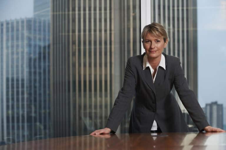 A woman in a business suit stands with hands on a conference table in a high-rise office, with tall buildings visible through the window behind her.
