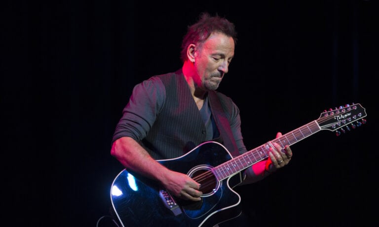 Bruce Springsteen plays a black acoustic guitar on stage under red and blue lighting, looking down with a focused expression.