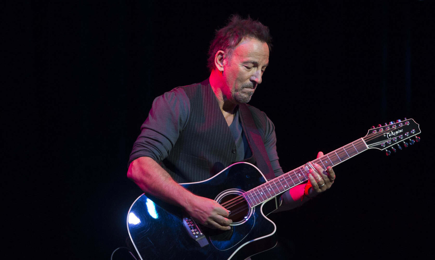 Bruce Springsteen plays a black acoustic guitar on stage under red and blue lighting, looking down with a focused expression.