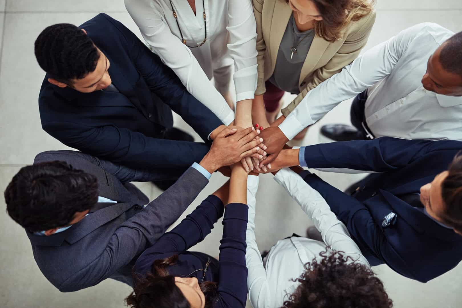 Group of business professionals standing in a circle with their hands stacked together in the center, viewed from above.