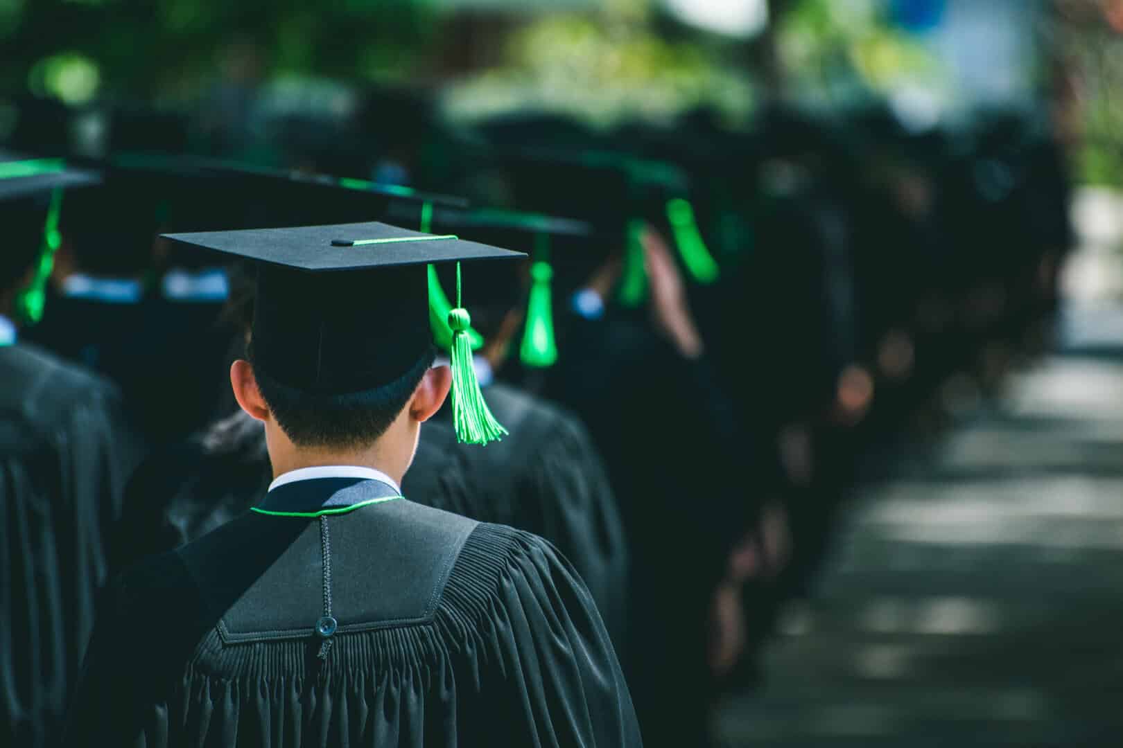 A group of graduates in black caps and gowns with green tassels stand in line outdoors, viewed from behind.