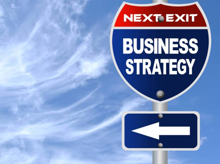 Highway sign reads “Next Exit Business Strategy,” with an arrow pointing left, set against a blue sky with wispy clouds.